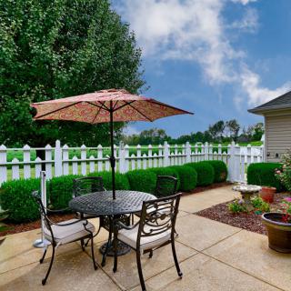 A cozy backyard patio with a table, chairs, and an umbrella next to a lush garden and white picket fence.