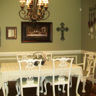 A formal dining room with a chandelier, ornate white table and chairs, decorated with wall art and a cross on green walls.