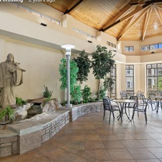 A serene indoor space featuring a statue and fountain, surrounded by greenery and a seating area under an arched wooden ceiling.