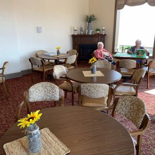 A cozy seating area with several tables and chairs, decorated with sunflowers in vases, while two people sit near a window.