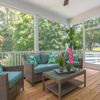 A cozy screened porch with wicker furniture, cushions, and a potted plant, overlooking lush greenery.