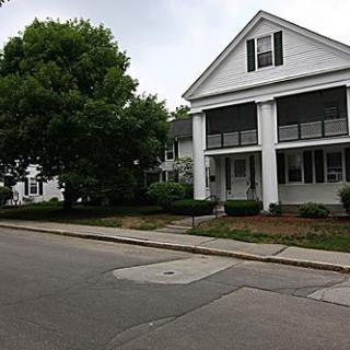 A view of a residential street with a large white house and surrounding trees.