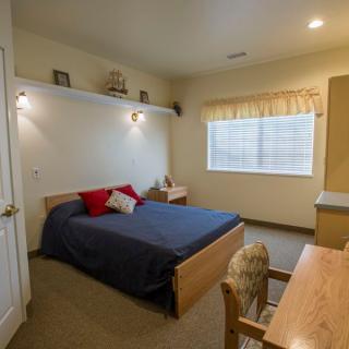 A cozy bedroom featuring a bed with blue bedding, a wooden desk, and a window with light curtains.