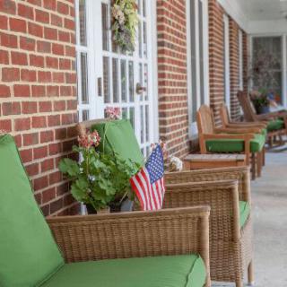 A row of wicker chairs with green cushions on a brick porch, featuring potted plants and small American flags.