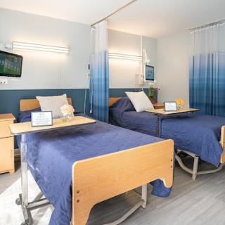 A modern patient room in a hospital featuring two beds with blue bedding and a television.