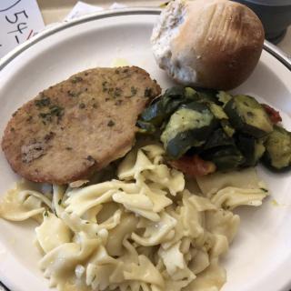 A meal featuring a slice of meat, pasta, zucchini, and a bread roll on a plate.