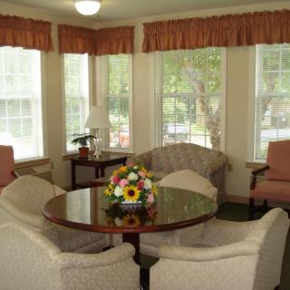 A cozy living room with a round table and comfortable chairs surrounded by large windows with light curtains.