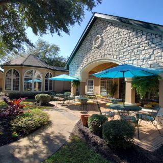 A charming outdoor patio area with tables and umbrellas, surrounded by lush garden landscaping and architectural features under a clear blue sky.