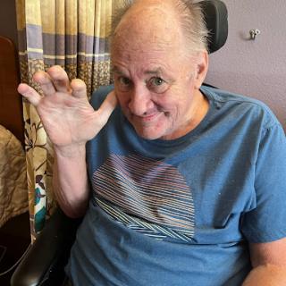 A smiling man in a wheelchair waving his hand indoors.