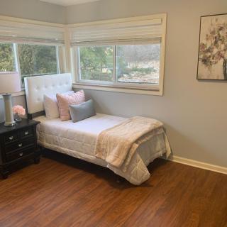 A cozy bedroom with a single bed beside large windows that allow natural light to flood the space, complete with a nightstand and decorative accents.