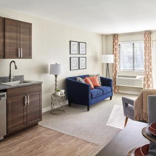 A modern apartment living area with a kitchenette, blue sofa, and large window.