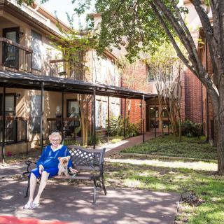 An elderly woman sits on a bench in a garden in front of a building.