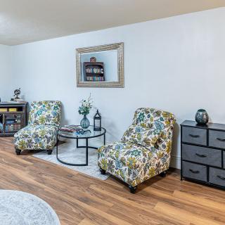 A cozy living room setup featuring patterned chairs, a glass table, and decor elements like a mirror and drawers against a wooden floor.