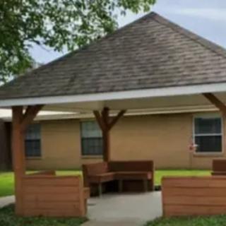 A gazebo with wooden benches located near a brick building, providing a shaded outdoor seating area.