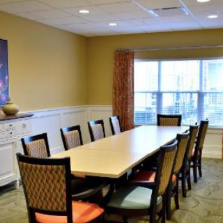 A conference room with a long meeting table and chairs, featuring large windows and bright lighting.