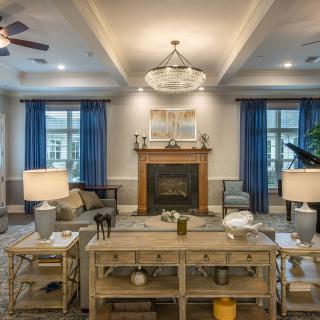 A cozy living room featuring a central fireplace, elegant chandelier, and stylish furniture with blue curtains framing large windows.