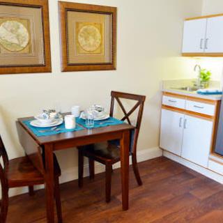 A cozy dining area with a small wooden table set for two, adjacent to a compact kitchenette, decorated with wall art.