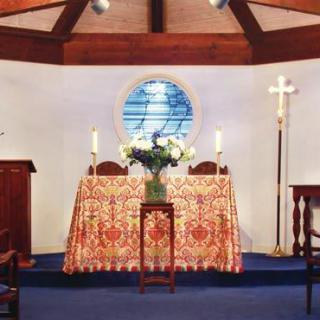 A church altar decorated with flowers, candles, and a cross, featuring a stained glass window in the background.