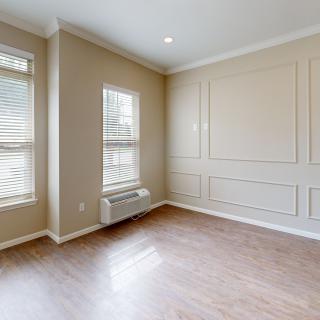 A well-lit empty room with hardwood flooring, beige walls, and two large windows letting in natural light.