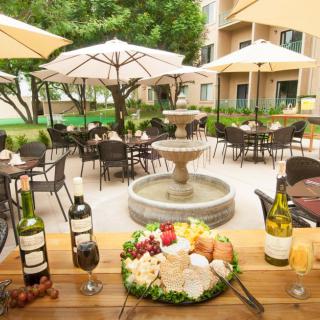 A charming outdoor restaurant patio with tables, chairs, and umbrellas surrounding a central fountain. A wooden table in the foreground is adorned with wine bottles and a cheese platter.