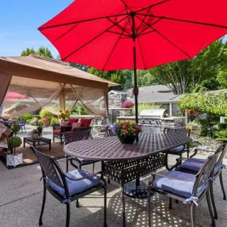 A cozy outdoor patio setup with a red umbrella shading a table and chairs, surrounded by a lush garden.