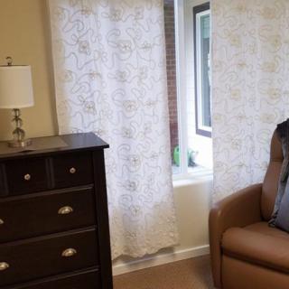 A cozy room with a dresser, armchair, lamp, and decorative curtains by the window.