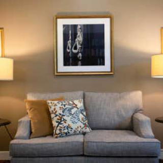 A cozy living room setup with a gray sofa, decorative pillows, and warm lighting from two lamps. A framed piece of art hangs above the sofa.