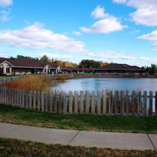 A scenic view of a pond surrounded by houses, with a fence and sidewalk in the foreground under a partly cloudy sky.