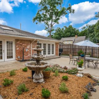 A charming backyard with a garden and fountain, featuring outdoor seating on a patio next to a brick house.