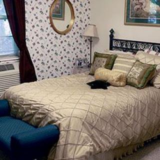 A cozy bedroom with patterned wallpaper, a neatly made bed, decorative pillows, and an elegant mirror on the wall.