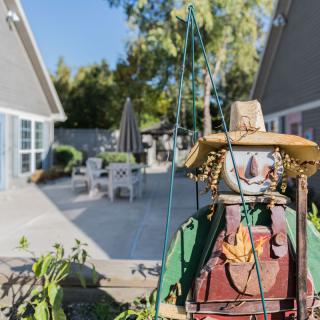 A decorative scarecrow in a sunny backyard garden with patio furniture in the background.