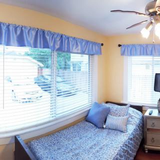 A cozy bedroom with a single bed, blue curtains, and yellow walls, illuminated by natural light from large windows.