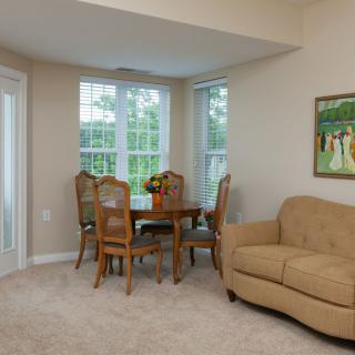 A cozy living room with a dining area featuring a sofa, round table with chairs, large windows, and a painting on the wall.
