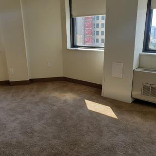 An empty room with carpeted flooring, featuring large windows letting in sunlight and an air conditioning unit.