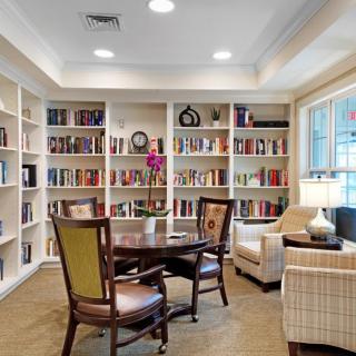 A cozy library room featuring bookshelves, a table with chairs, and a lamp by the window.