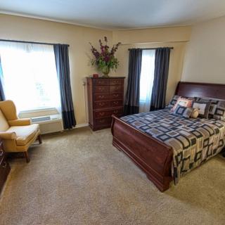 A well-furnished bedroom with a bed, dresser, armchair, and large windows.