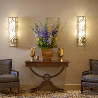 An elegantly decorated interior with a central table featuring a vase of flowers, flanked by two chairs and illuminated by wall sconces.