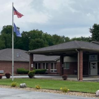 A brick building with a covered entrance, accompanied by a flagpole displaying an American flag, set against a backdrop of trees and a cloudy sky.