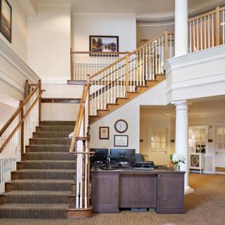 A well-lit interior space featuring a staircase with wooden railings, a reception desk, and decorative columns.