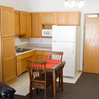 A compact kitchen in an apartment featuring wooden cabinets, a white refrigerator, a microwave, and a small dining table with chairs.