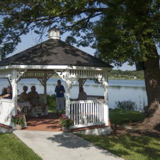 A group of people sitting in a gazebo by the lake, surrounded by trees and picturesque scenery.