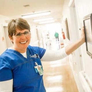 A smiling nurse in a blue uniform stands in a hospital corridor.