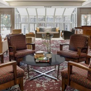 A spacious lobby with comfortable seating and a central table, featuring large windows in the background.