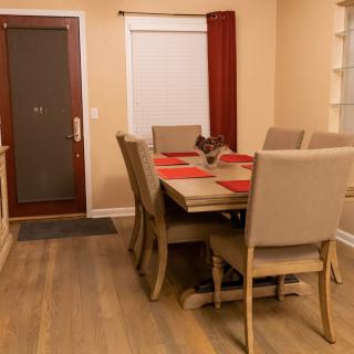 A cozy dining room featuring a wooden table set with red placemats, surrounded by upholstered chairs on a wooden floor.