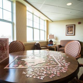 A cozy lounge area with a puzzle in progress on the table, surrounded by chairs and bathed in natural light from large windows.