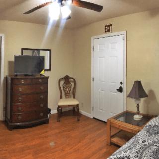 A cozy room featuring a wooden dresser with a TV on top, a classic chair, and a small table with a lamp, all set against warm-toned walls and hardwood flooring.