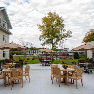 A charming outdoor patio with wooden tables and chairs, shaded by umbrellas, set amidst a garden with a large tree and buildings in the background under a cloudy sky.