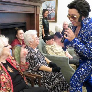 A performer in a blue costume sings to an elderly audience indoors.