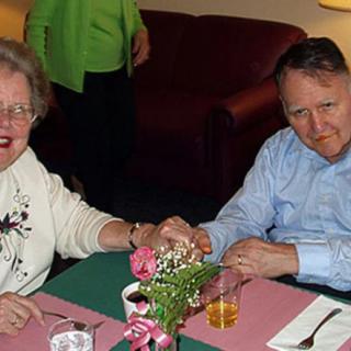 An elderly couple holding hands and smiling at a dining table, decorated with a floral arrangement and drinks.