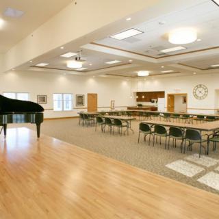 A spacious event hall with a piano on stage and several tables and chairs arranged for a meeting or gathering.